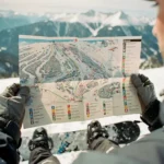 Snowboarder hält Pistenplan in behandschuhten Händen vor verschneiter Bergkulisse, Snowboard-Boots sichtbar