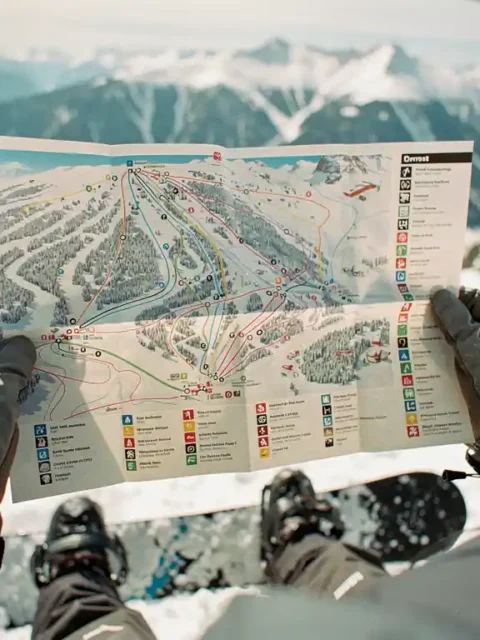 Snowboarder hält Pistenplan in behandschuhten Händen vor verschneiter Bergkulisse, Snowboard-Boots sichtbar