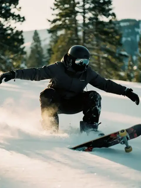 Snowboarder in donkere winterkleding gehurkt op poedersneeuw met skateboard-geïnspireerde houding op bergpiste