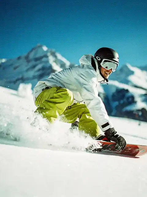 Snowboarder in felle winterkleding maakt bocht in poedersneeuw met bergtoppen op achtergrond onder blauwe hemel