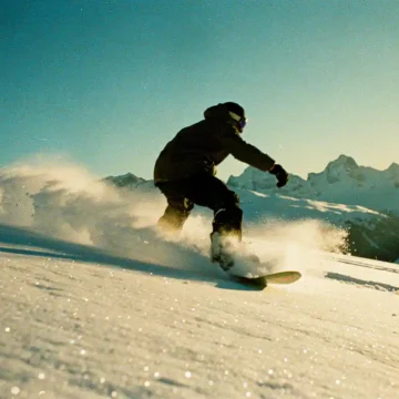 Snowboarder maakt bocht door poedersneeuw op bergpiste met alpine toppen en blauwe lucht op achtergrond