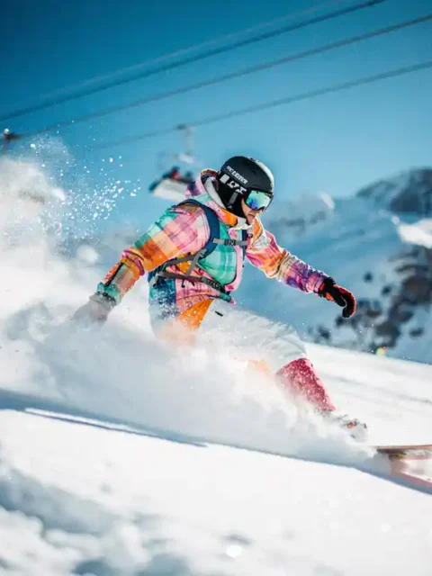 Snowboarder in felgekleurde winterkleding carvet door poedersneeuw op zonnige bergpiste met skilift op achtergrond