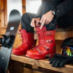 Snowboarder past rode snowboardlaarzen aan op houten bank in alpine lodge met snowboard en uitrusting eromheen