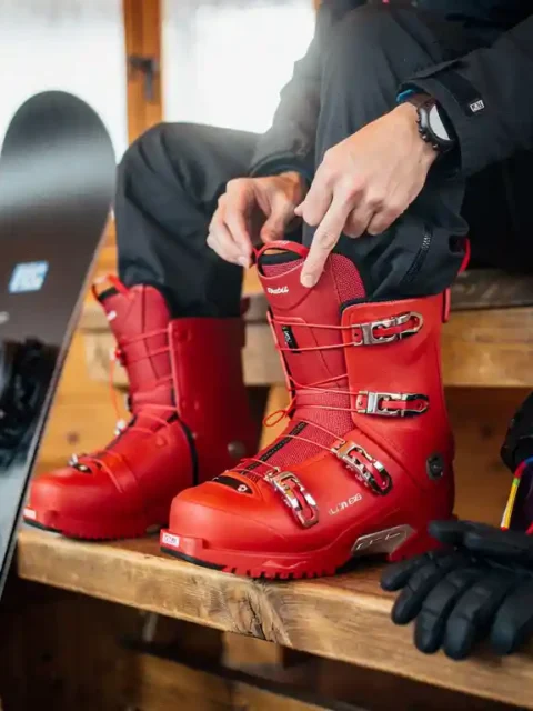 Snowboarder past rode snowboardlaarzen aan op houten bank in alpine lodge met snowboard en uitrusting eromheen