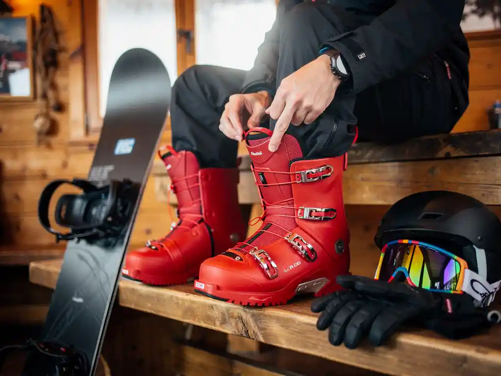 Snowboarder past rode snowboardlaarzen aan op houten bank in alpine lodge met snowboard en uitrusting eromheen