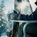Snowboarder's gloved hands gripping colorful snowboard on ski lift chair with snowy pine trees below