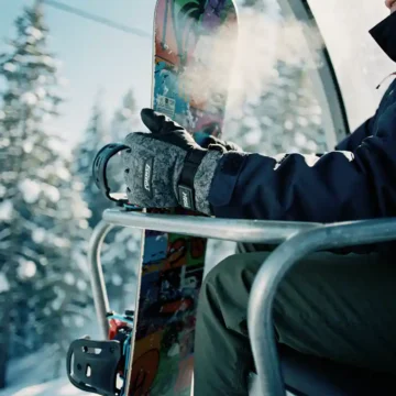 Snowboarder's gloved hands gripping colorful snowboard on ski lift chair with snowy pine trees below