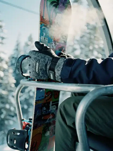 Snowboarder's gloved hands gripping colorful snowboard on ski lift chair with snowy pine trees below