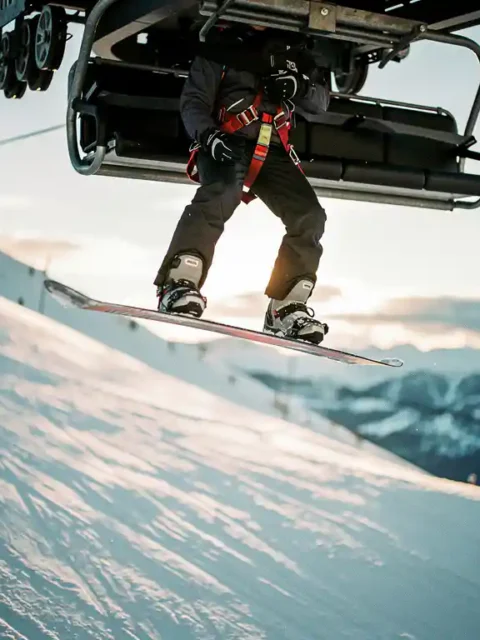 Snowboarder suspended mid-air beneath ski lift chair with safety harness, snow-covered mountain slope below in afternoon light.