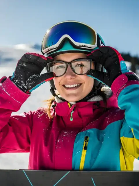 Snowboarder met beschermende bril en glimlach op besneeuwde bergpiste met dennenbomen en alpine zonnestralen