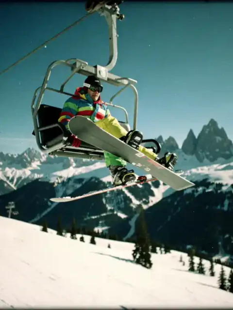 Snowboarder hängt hilflos am Skilift über verschneiter Berglandschaft, ein Fuß teilweise aus der Bindung gerutscht