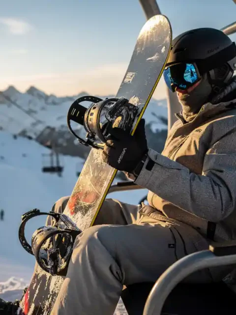 Snowboarder op skilift houdt snowboard verticaal vast, besneeuwde berghellingen op achtergrond