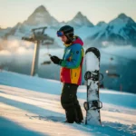 Snowboarder in felle winterkleding controleert smartphone op besneeuwde piste met bergtoppen op achtergrond