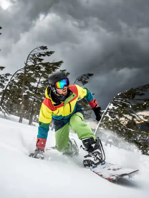 Snowboarder in felgekleurde winterkleding kijkt naar donkere stormwolken op besneeuwde bergpiste met buigende dennenbomen