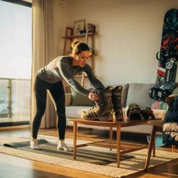Atleet doet dynamische rekoefeningen in woonkamer, reikt naar snowboardlaarzen op houten tafel, warme zonnestralen