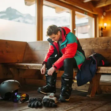 Beginnende snowboarder rijgt laarzen in skihut met uitgestalde winteruitrusting en besneeuwde bergen op achtergrond