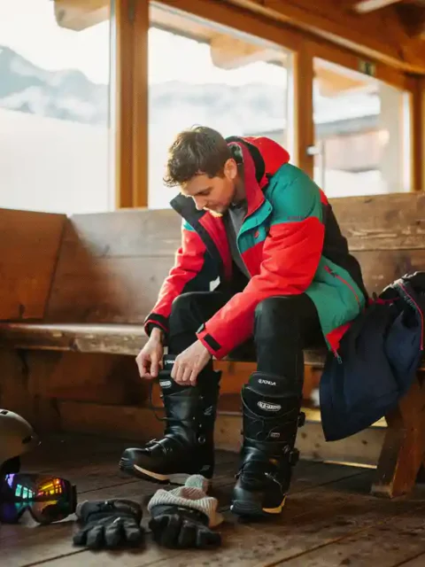 Beginnende snowboarder rijgt laarzen in skihut met uitgestalde winteruitrusting en besneeuwde bergen op achtergrond