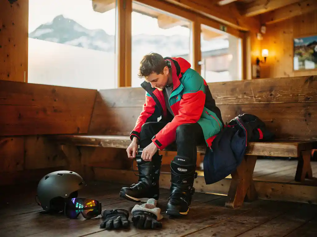 Beginnende snowboarder rijgt laarzen in skihut met uitgestalde winteruitrusting en besneeuwde bergen op achtergrond