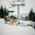 Snowboarder in felle winterkleding ligt in poedersneeuw onder skilift, ziet er opgelucht uit na val