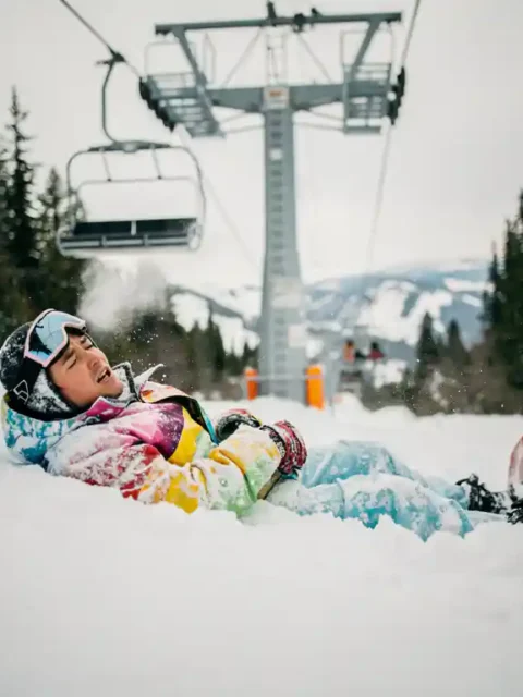 Snowboarder in felle winterkleding ligt in poedersneeuw onder skilift, ziet er opgelucht uit na val