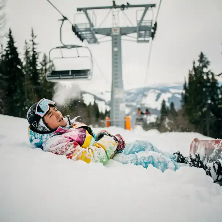 Snowboarder in felle winterkleding ligt in poedersneeuw onder skilift, ziet er opgelucht uit na val