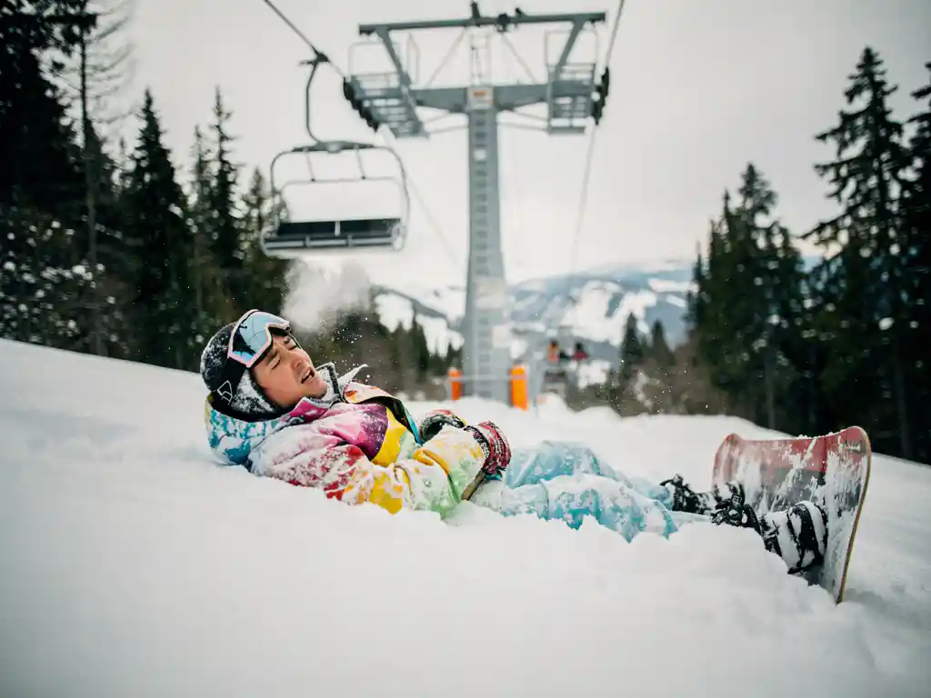 Snowboarder in felle winterkleding ligt in poedersneeuw onder skilift, ziet er opgelucht uit na val